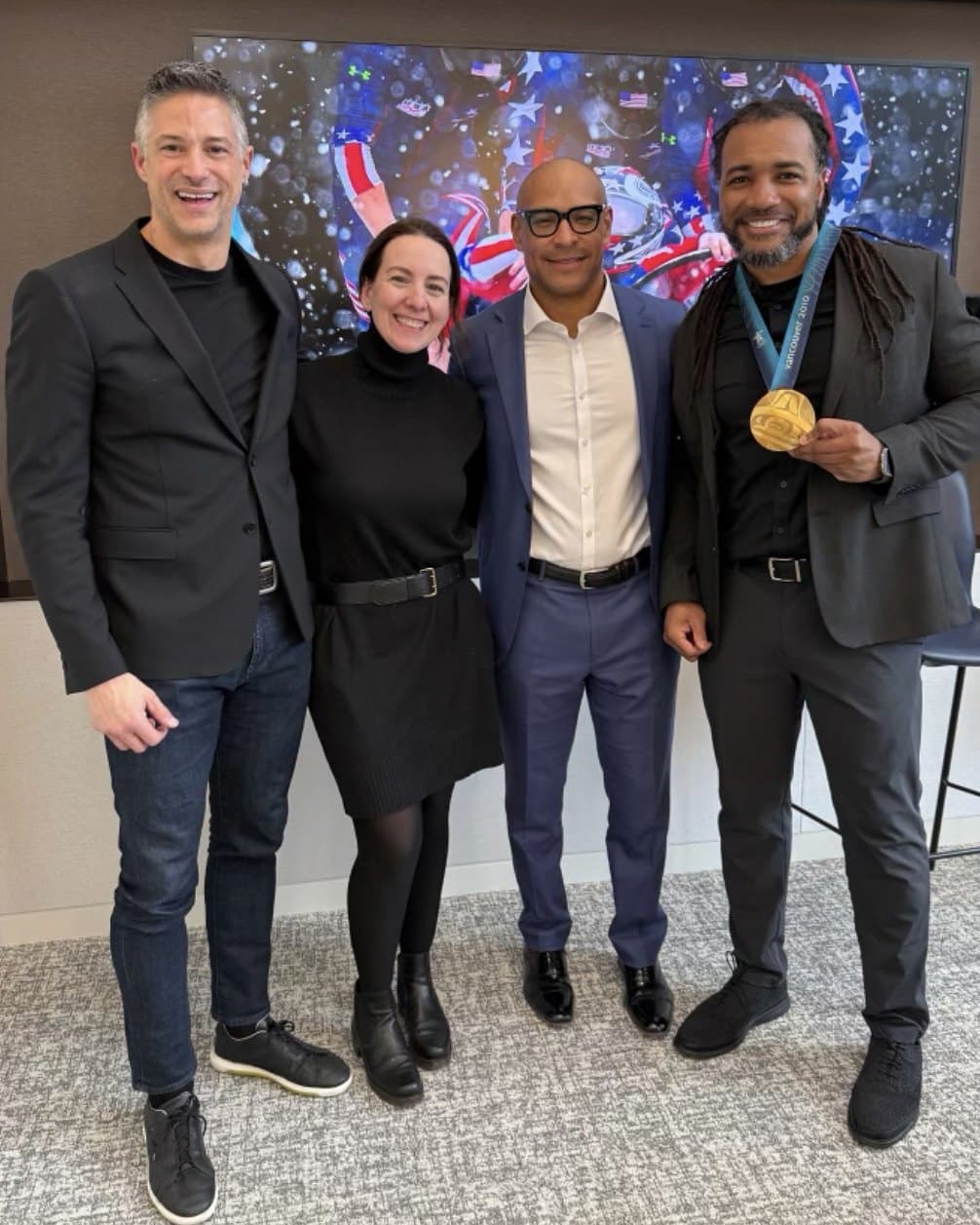 Usama Young, smiling, holding a gold medal, standing with colleagues in front of a screen